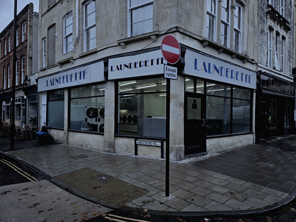 A 24-hour launderette on an ordinary UK high street, chosen because appliances that run themselves at 3am are already half-believed to be haunted before anything happens.