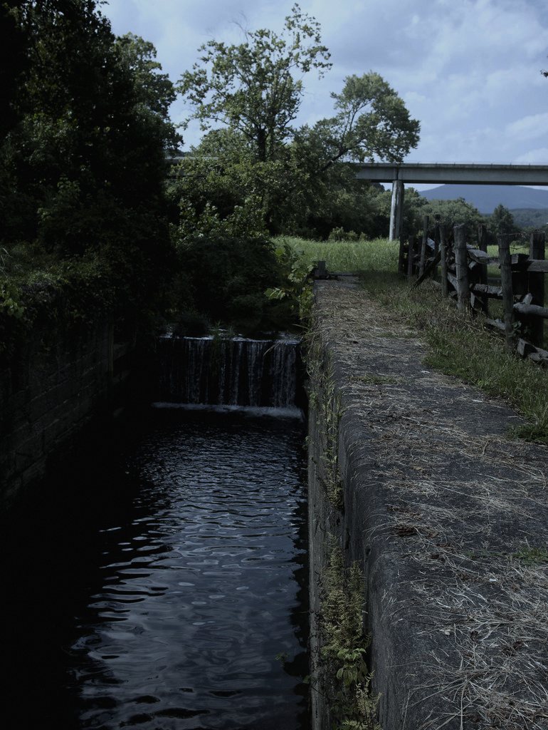 Set at a fictional canal lock in the English Midlands, chosen because lock-keeping still runs on paper logs where a stranger's handwriting would stand out immediately.